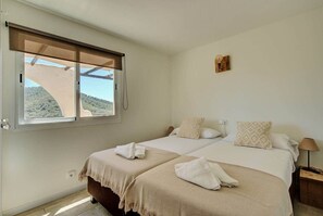 A bedroom with two beds, a window with a view of a hill, a small wall-mounted light, and a wooden nightstand. - Apartment nº7, 1 bedroom, 1 bathroom (Cala Llonga)
