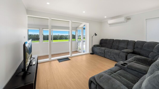 A modern living room with a sectional sofa, a flat-screen TV, and a large sliding glass door leading to a balcony with a view of greenery. - Lakeside 3 BDR House (Koonawarra)