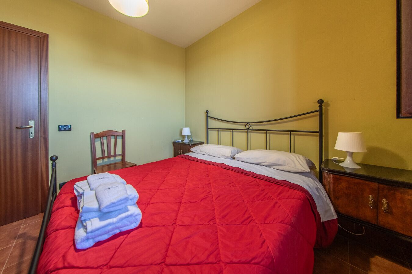 Een net opgemaakt bed met een rode quilt, een houten stoel, een nachtkastje met een lamp en een houten garderobe.