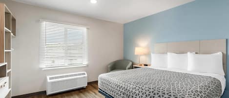A hotel room with a bed, a chair, a desk with a lamp, a window with blinds, and a built-in heater.