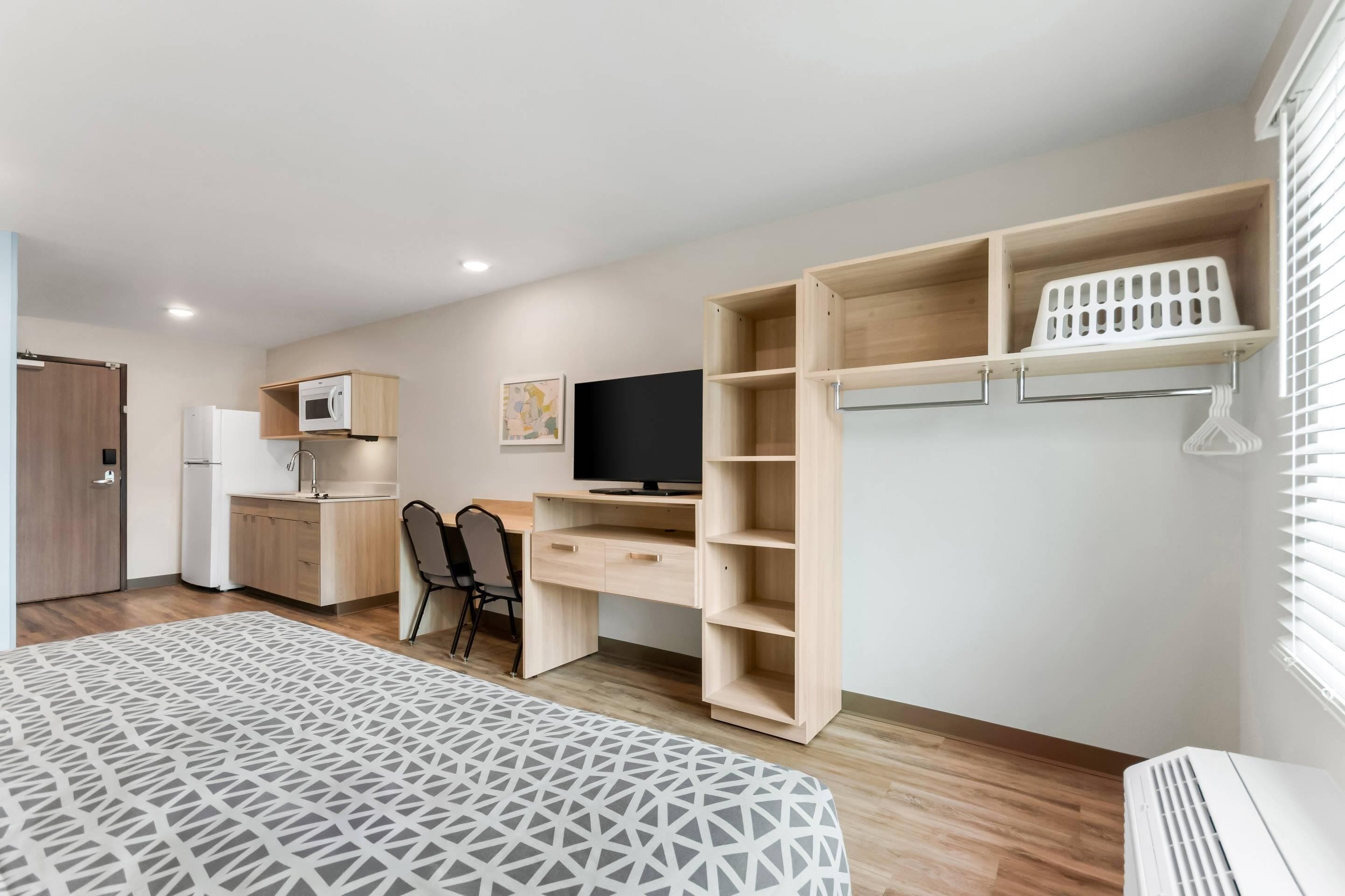 Una habitación moderna y bien iluminada con cama, escritorio, televisión, kitchenette y armario con estantes.