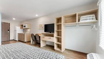 Une chambre moderne et bien éclairée, équipée d’un lit, d’un bureau, d’une télévision, d’une kitchenette et d’une armoire avec des étagères.
