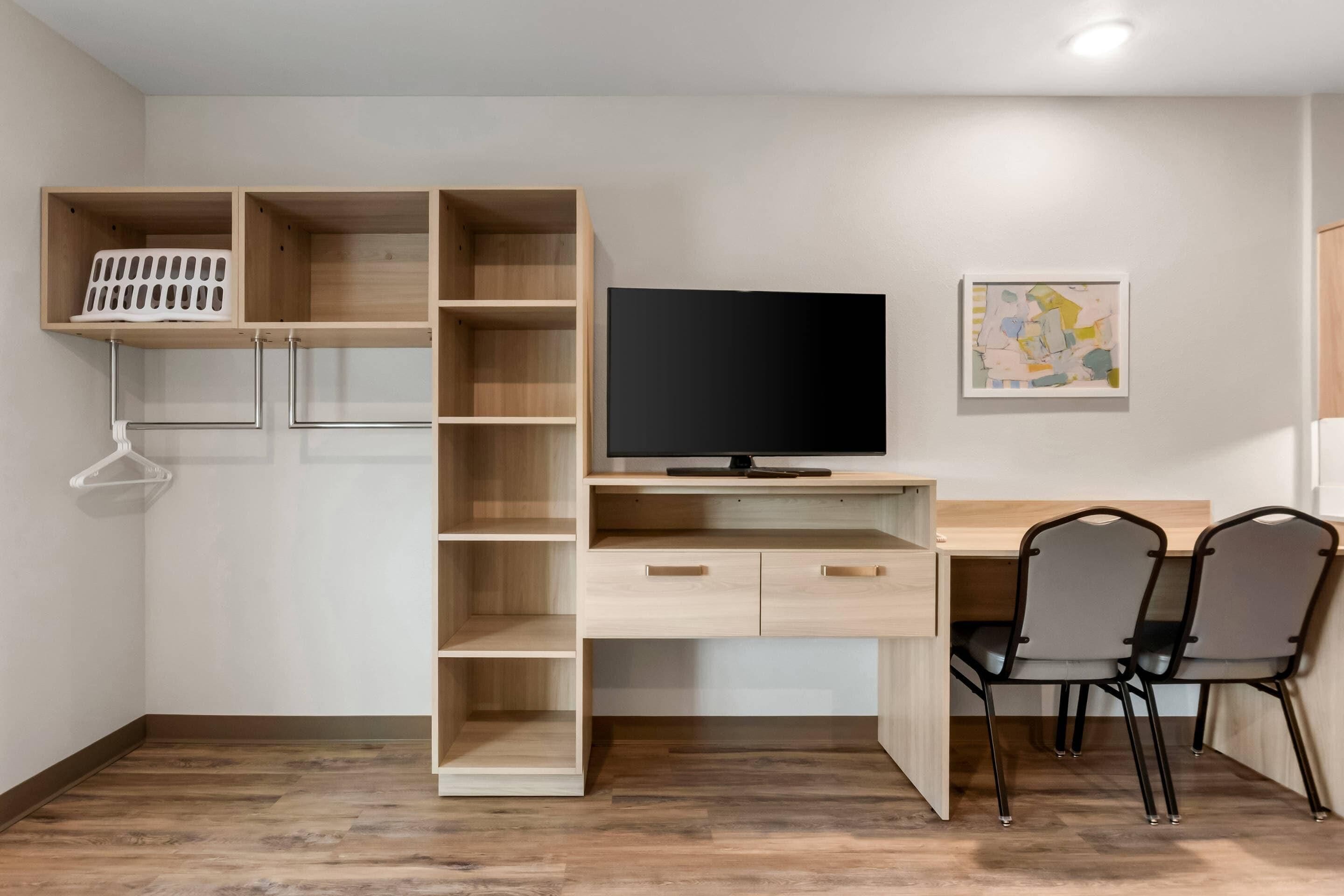 A room with a wooden desk, a television, two chairs, and a wall-mounted shelf with a laundry basket.