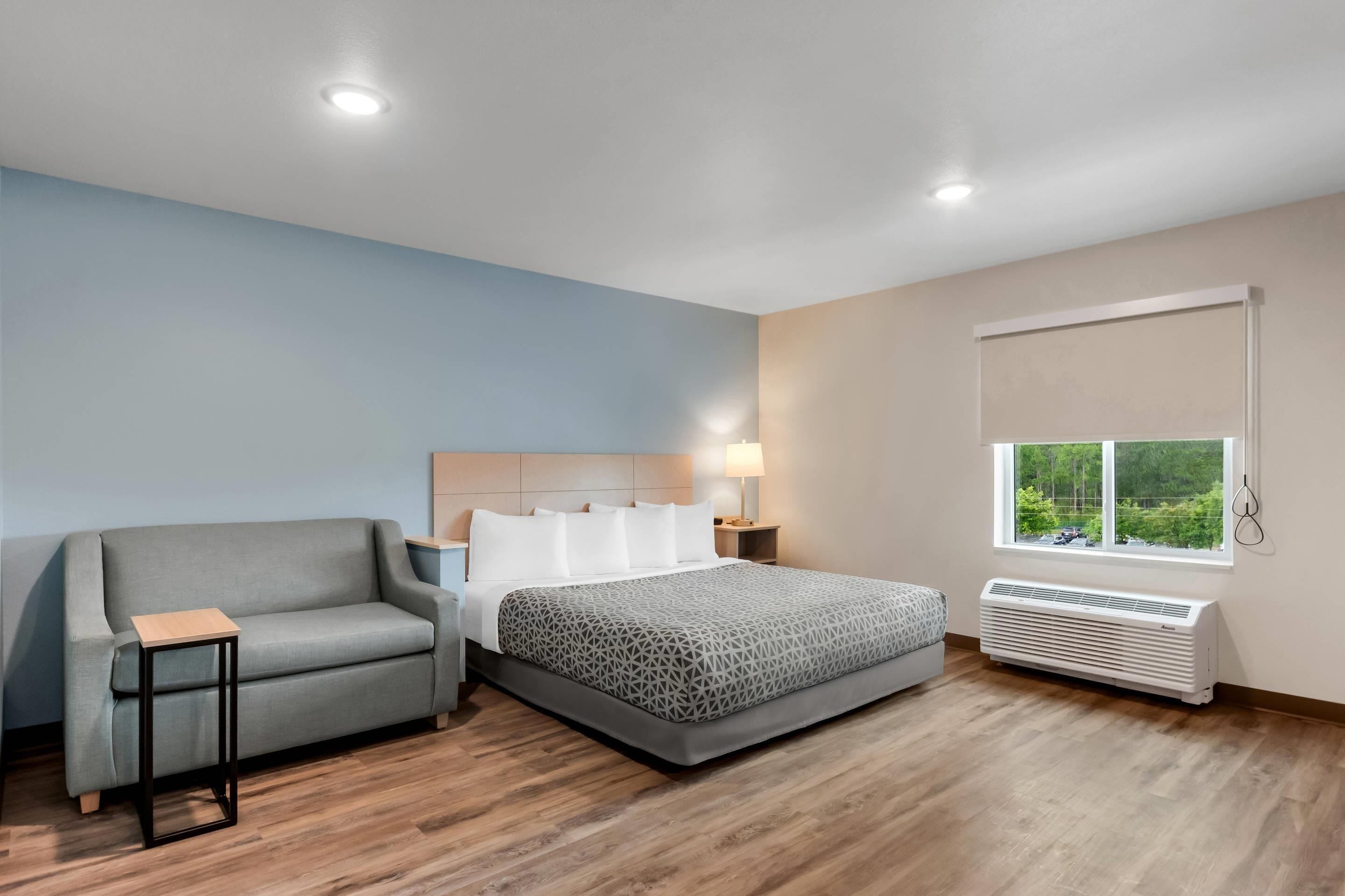 A bedroom with a bed, a sofa, a bedside table, a lamp, a window with a view, and a wall-mounted air conditioner.