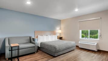 A bedroom with a bed, a sofa, a bedside table, a lamp, a window with a view, and a wall-mounted air conditioner.