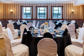 A banquet hall with round tables set for a formal event, large windows with a view of the water, and striped wallpaper.