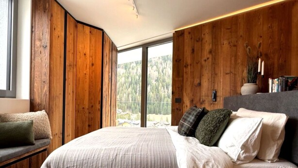 Une chambre avec un grand lit, des murs recouverts de panneaux de bois, une fenêtre donnant sur les arbres et une table de chevet avec des livres.