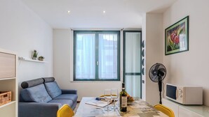 A compact living space with a dining table set for two, a sofa, a fan, a microwave, and a window with curtains. - Cosy apartment for 2 guests with WIFI, patio, pets allowed and panoramic view (Como)