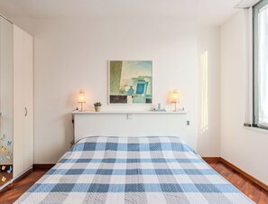 Ein Schlafzimmer mit kariertem Bettbezug, zwei Nachttischlampen, einem kleinen Topfpflanzen und einem Bild an der Wand.