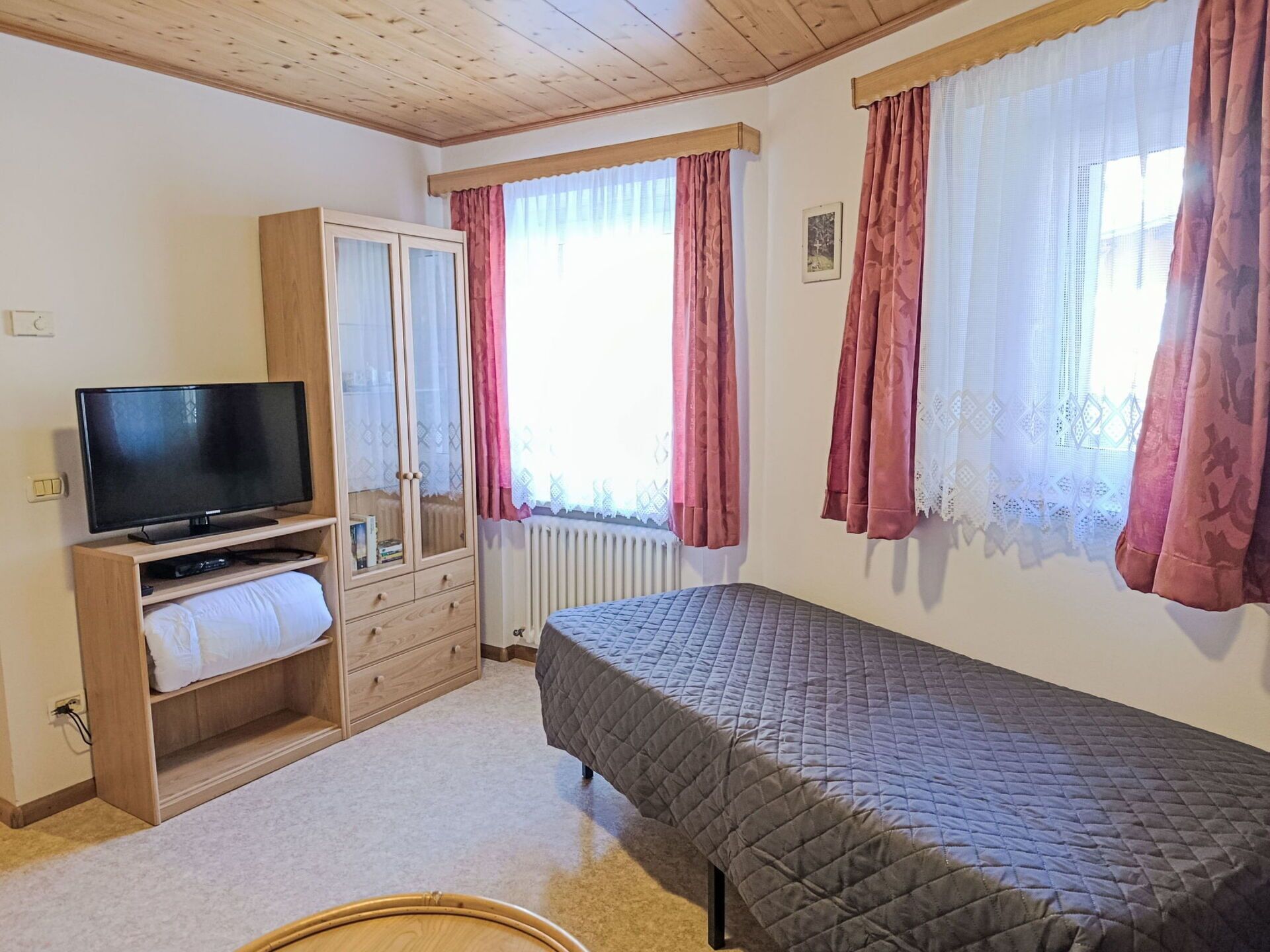 Une chambre à coucher avec un lit, une télévision, une armoire et une fenêtre avec des rideaux.