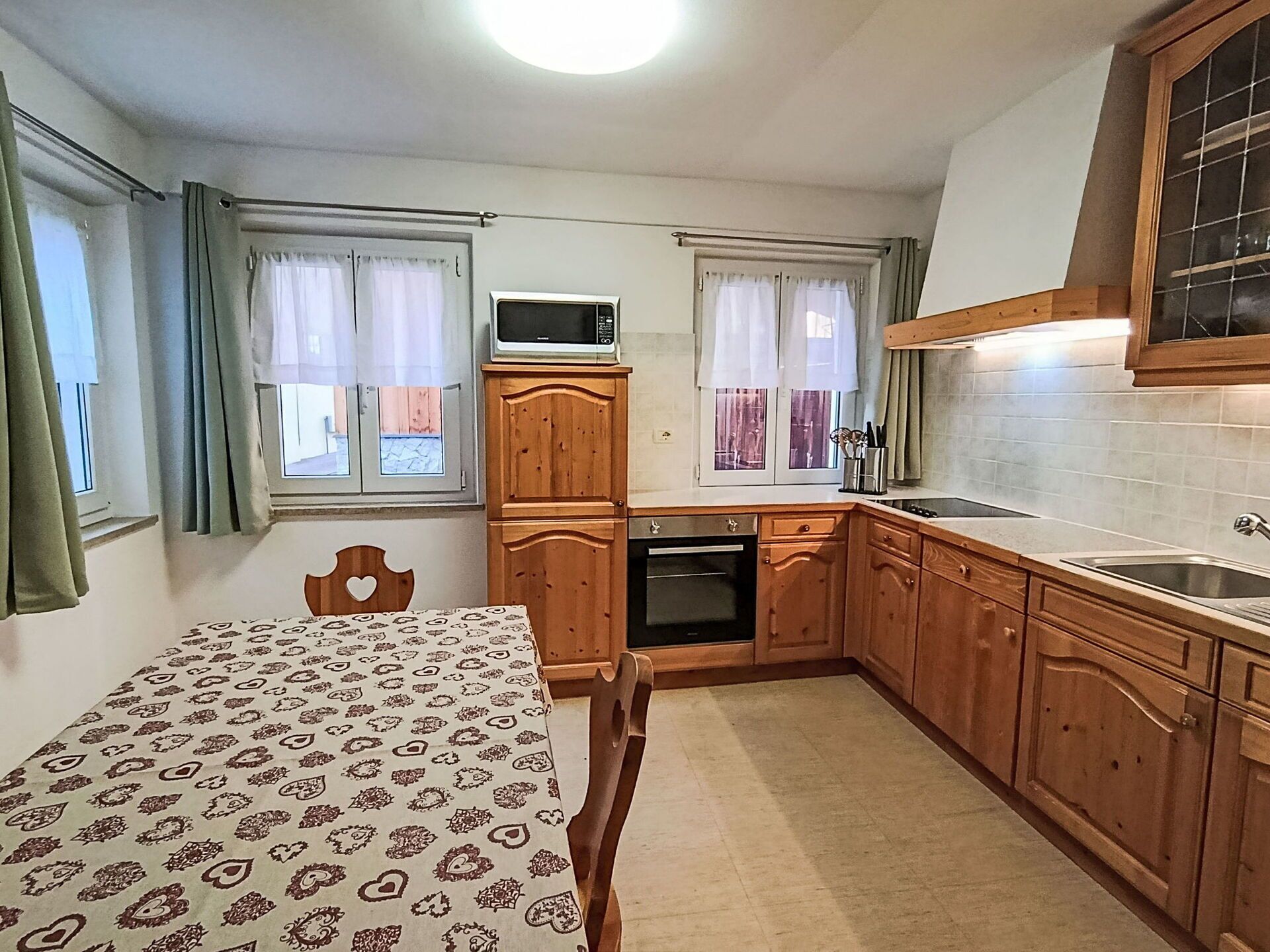 Cocina compacta con armarios de madera, microondas, pileta y comedor con mantel estampado.