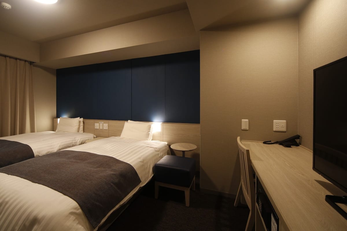 Ein Hotelzimmer mit zwei Betten, einem Schreibtisch mit Stuhl, einem Fernseher und einem kleinen Tisch.