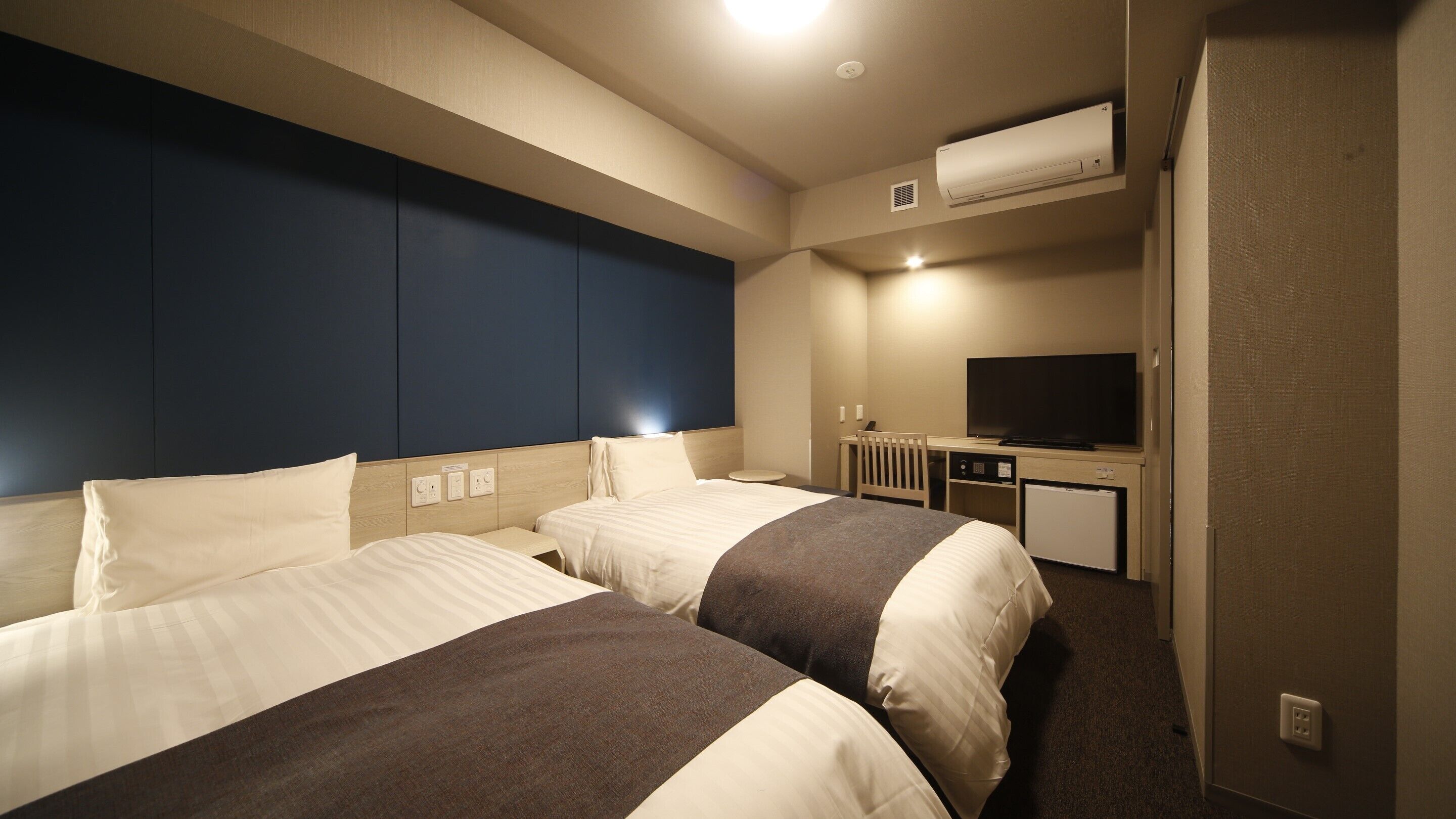 A hotel room with two beds, a desk, a television, and an air conditioning unit.