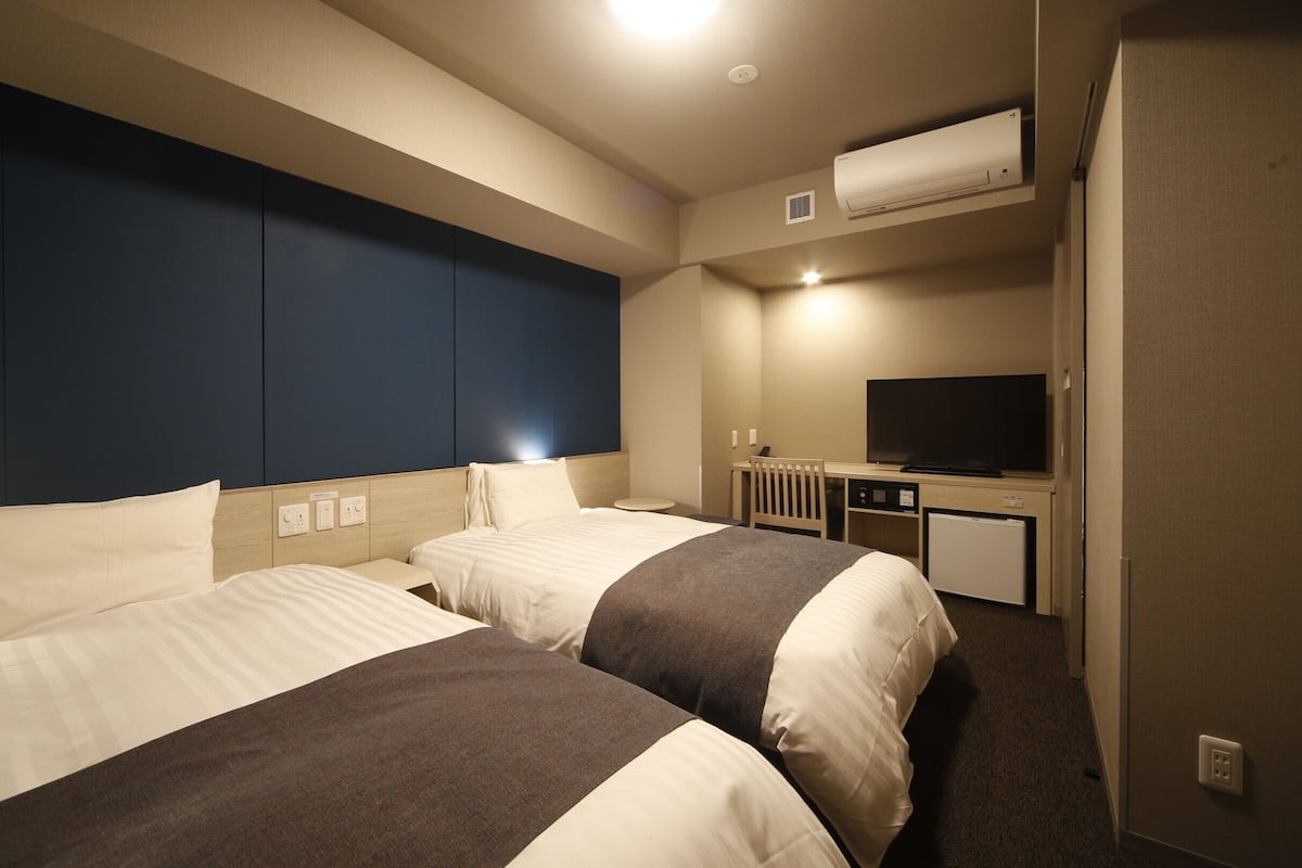 Ein Hotelzimmer mit zwei Betten, einem Schreibtisch, einem Fernseher und einer Klimaanlage.