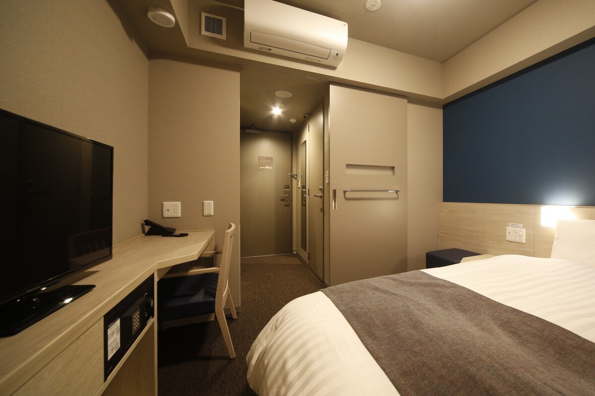 Quarto de hotel com cama, escrivaninha com televisão, porta e aparelho de ar condicionado.