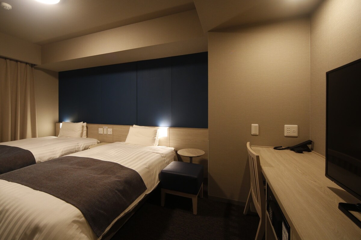 Quarto de hotel com duas camas, uma escrivaninha com cadeira, uma televisão e uma mesinha.