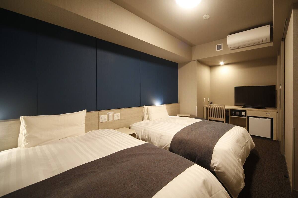 Ein Hotelzimmer mit zwei Betten, einem Fernseher, einer Minikühlschrank und einer Klimaanlage.