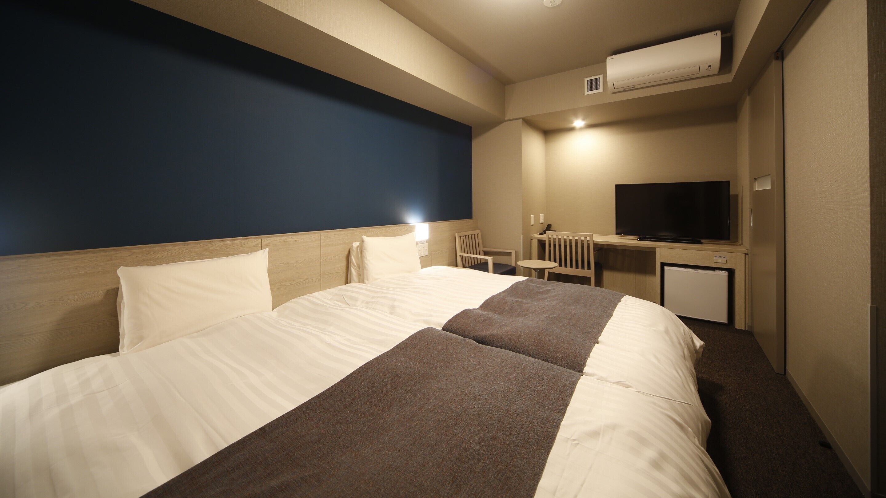 A hotel room with a bed, a desk, a chair, a television, and an air conditioning unit.