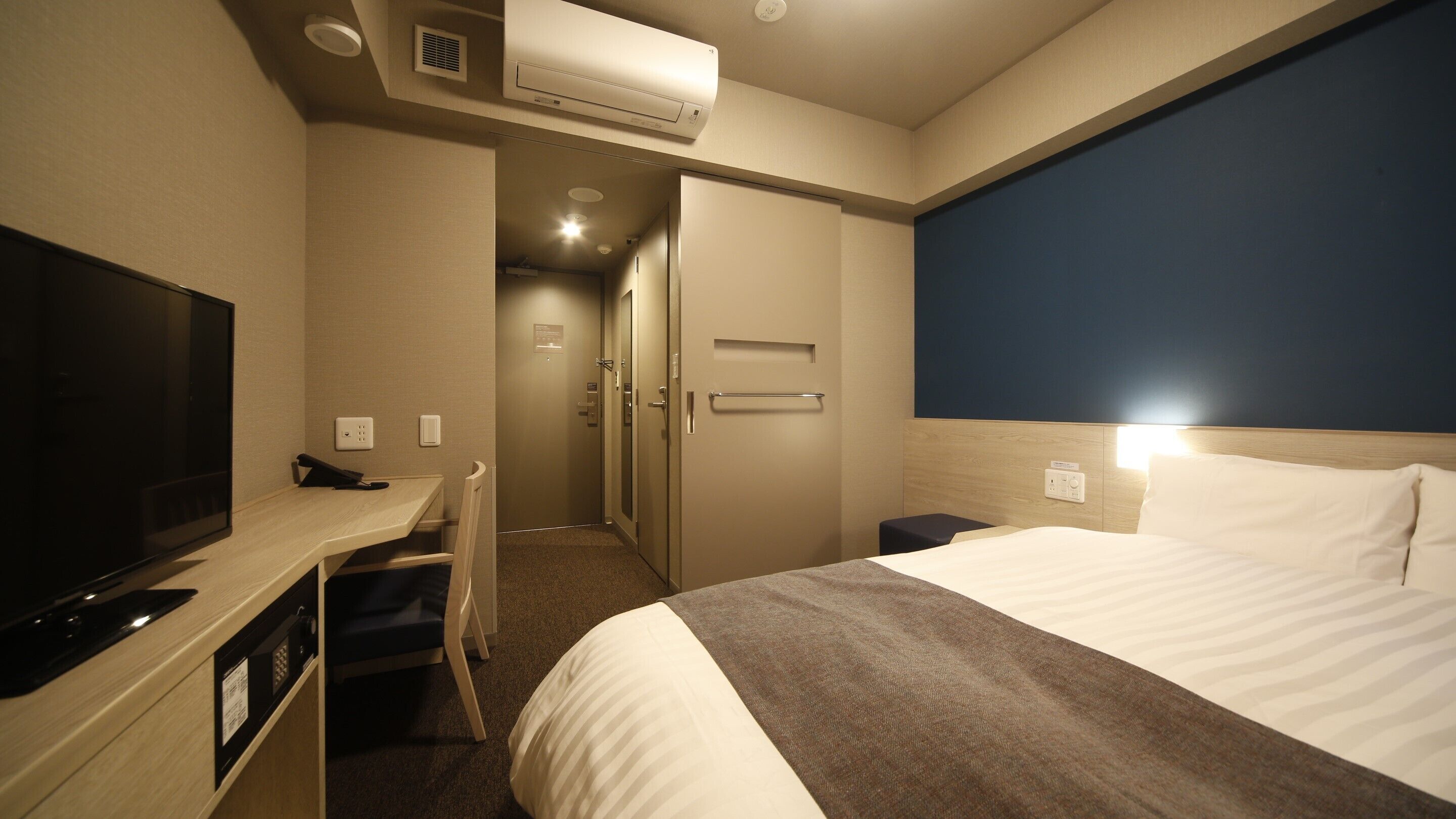 A hotel room with a bed, a desk with a television, a door, and an air conditioning unit.