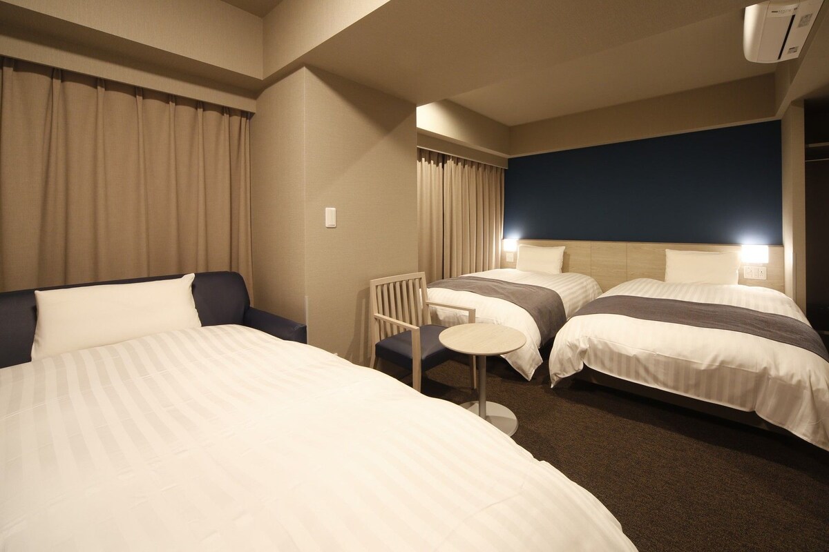Quarto de hotel com duas camas, uma cadeira, uma mesinha e um guarda-roupa.