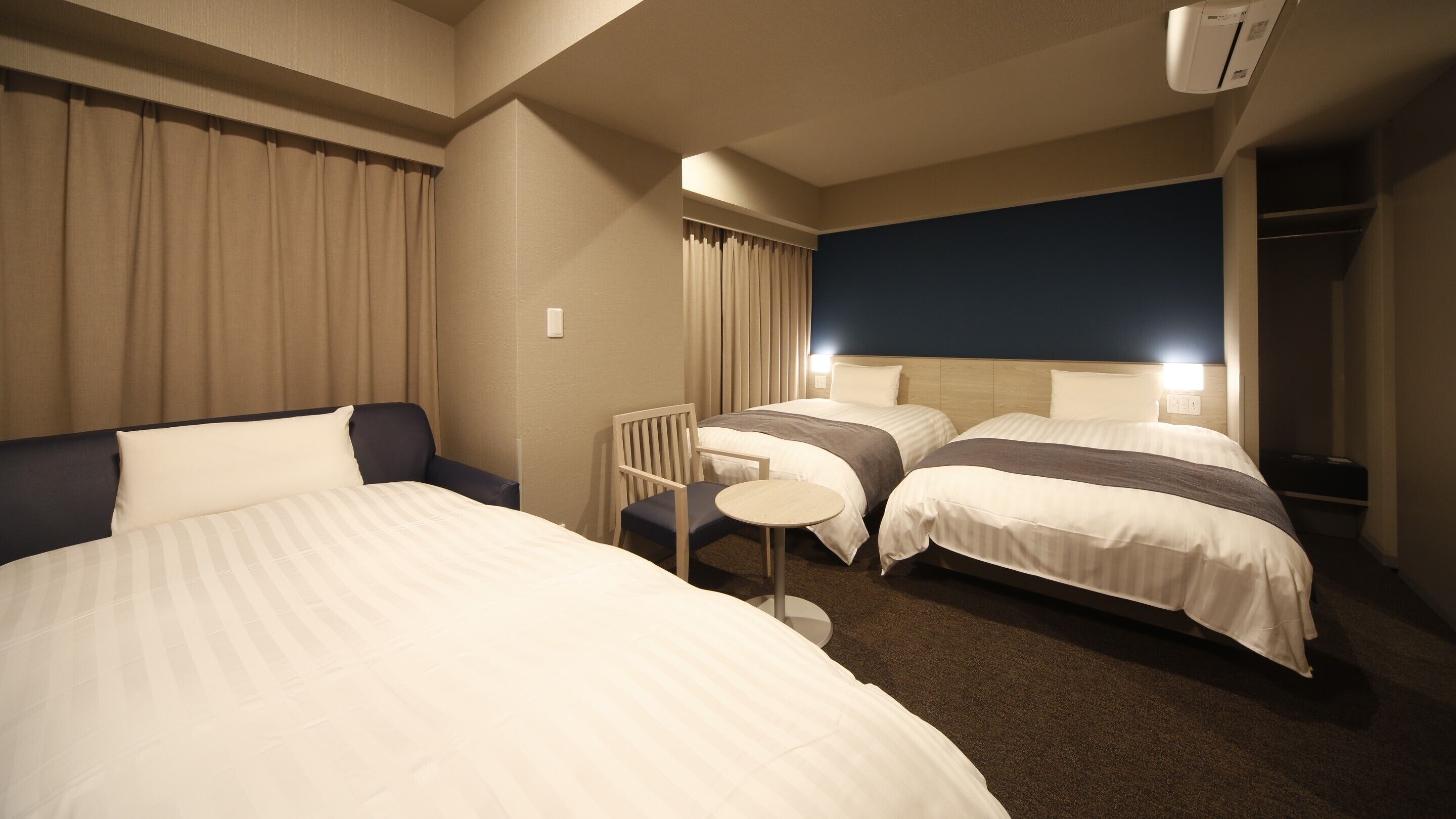 A hotel room with two beds, a small table, a chair, a ceiling fan, and a wardrobe.