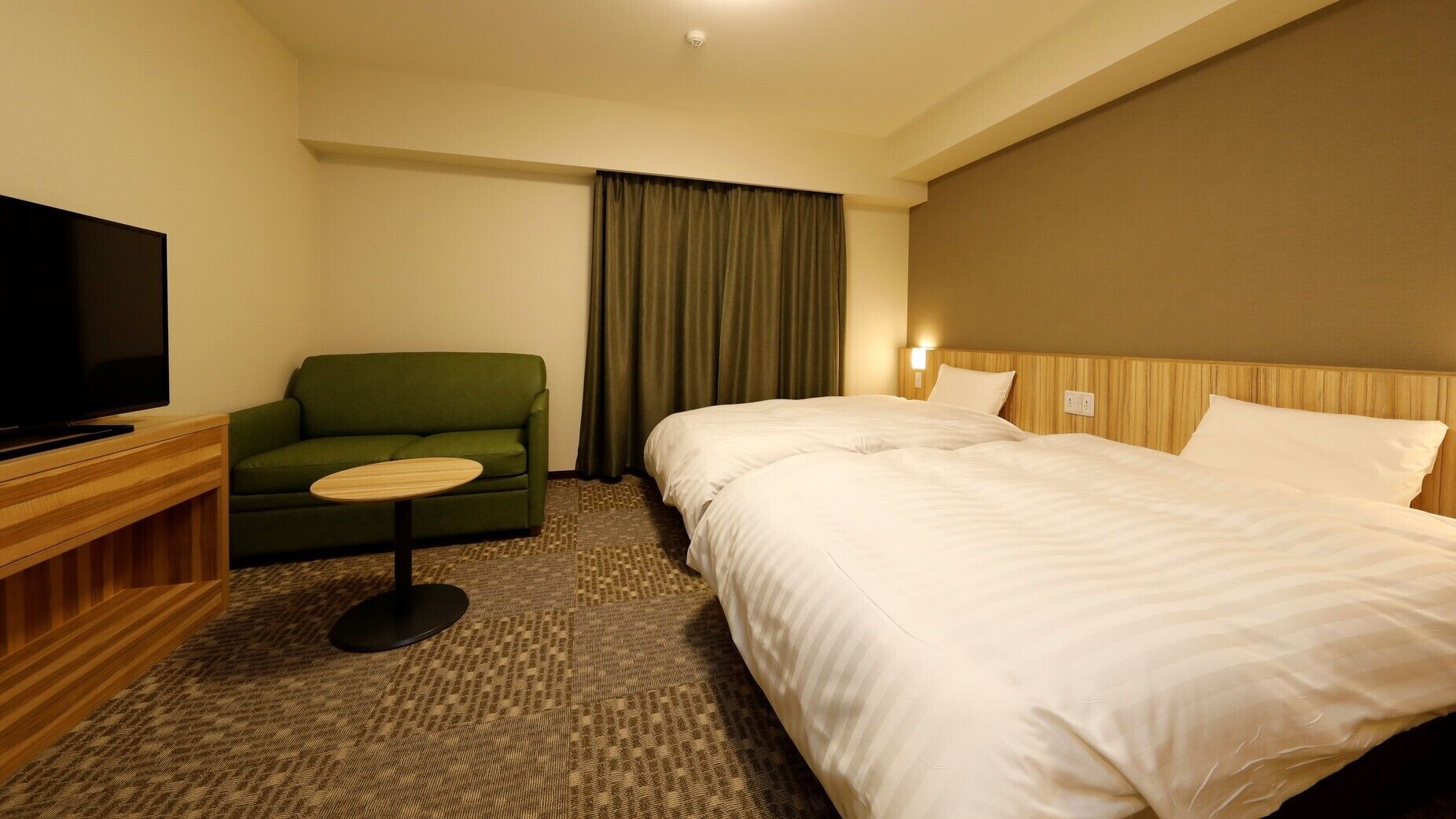 A hotel room with a bed, a television, a green armchair, and a small table.