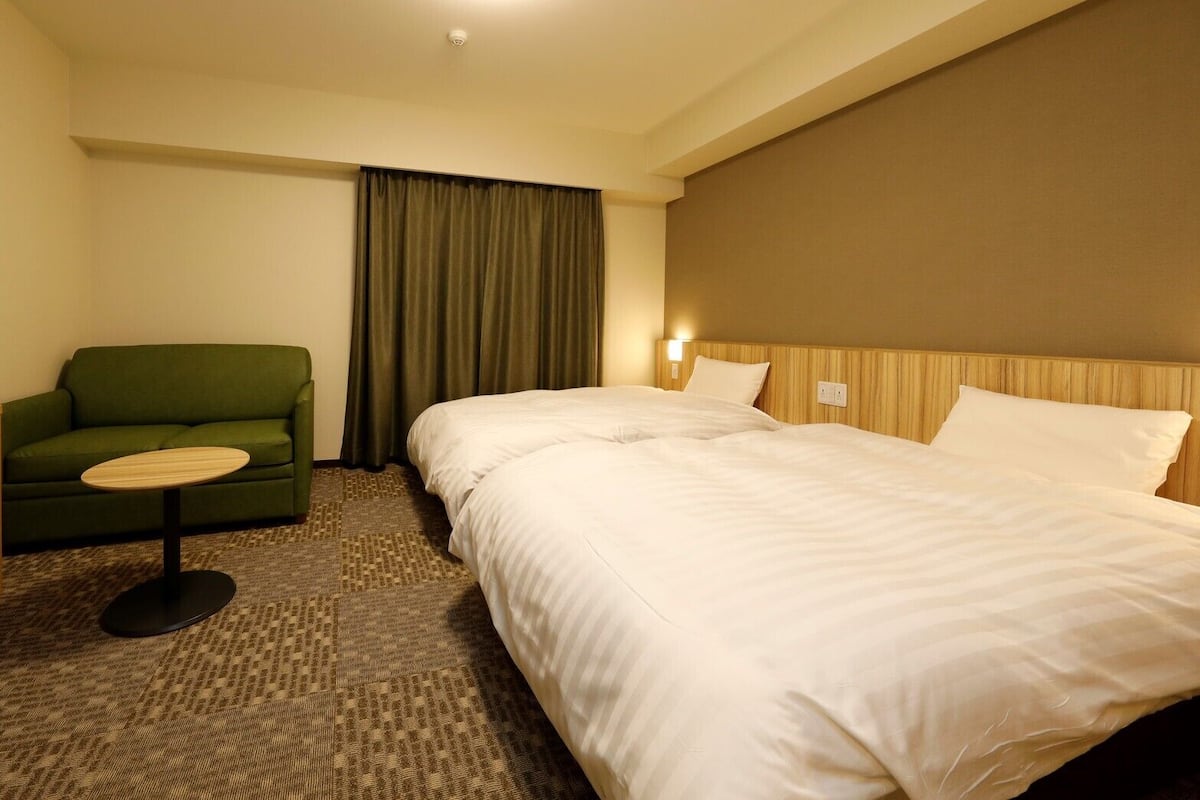 Una habitación de hotel con una cama, televisión, un sillón verde y una mesita.