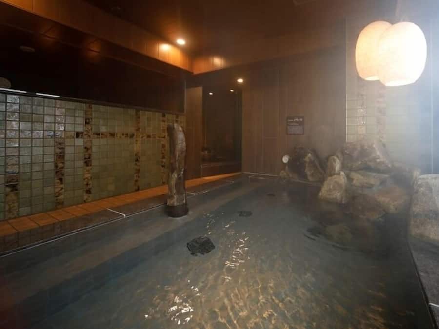 A hot spring bath with tiled walls, a wooden floor, and a stone wall with rocks.