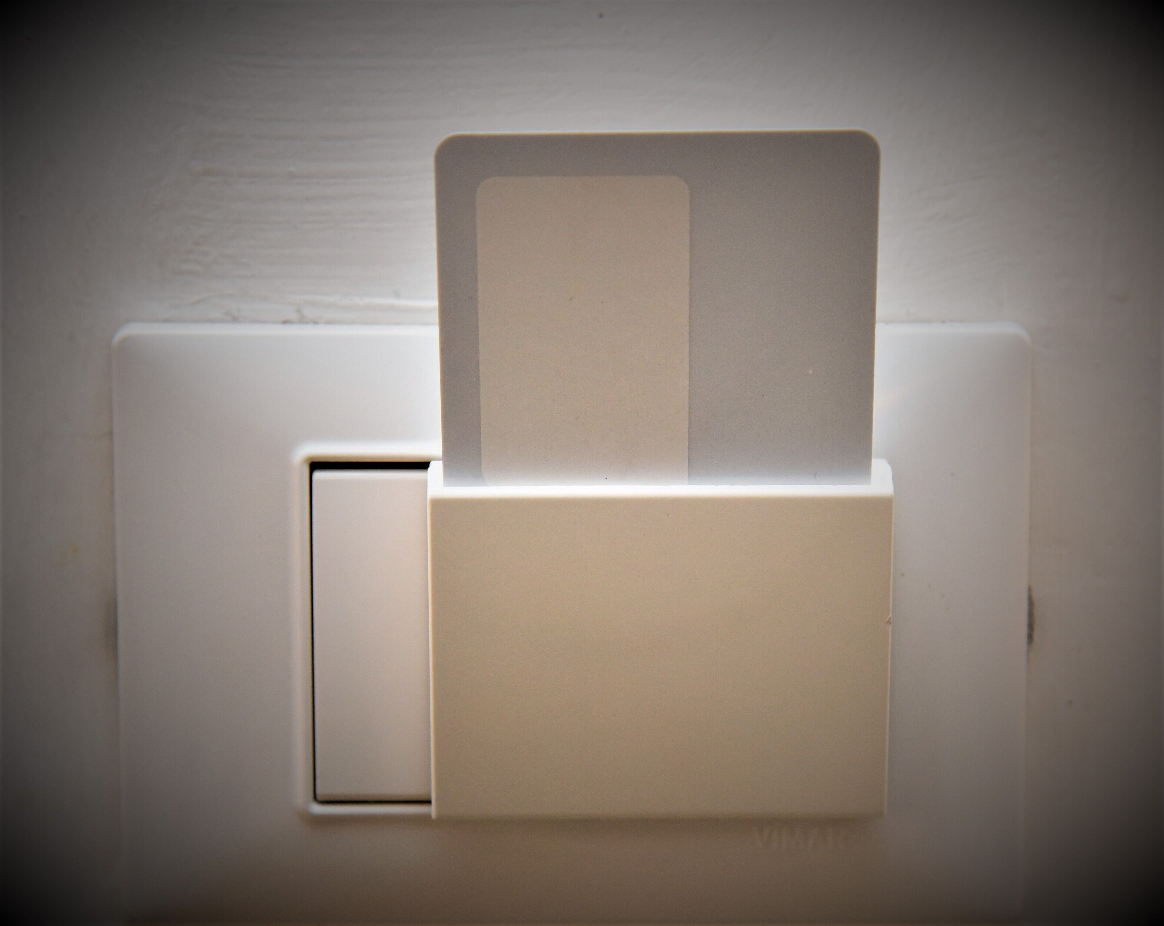 Pared blanca con un interruptor de luz y un objeto rectangular, quizás una tarjeta o una etiqueta, insertado en una ranura.