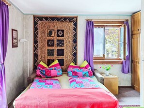 Ein Schlafzimmer mit Bett, Fenster, Wandteppich und einem kleinen Tisch mit Vase.