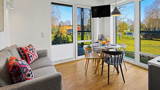 Ein modernes Wohnzimmer mit Sofa, Esstisch mit Stühlen, Fernseher und großen Fenstern mit Blick ins Freie.