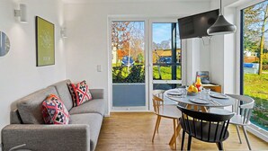 Ein modernes Wohnzimmer mit grauem Sofa, einem Esstisch mit Stühlen, einem Fernseher und einem großen Fenster mit Außenblick.