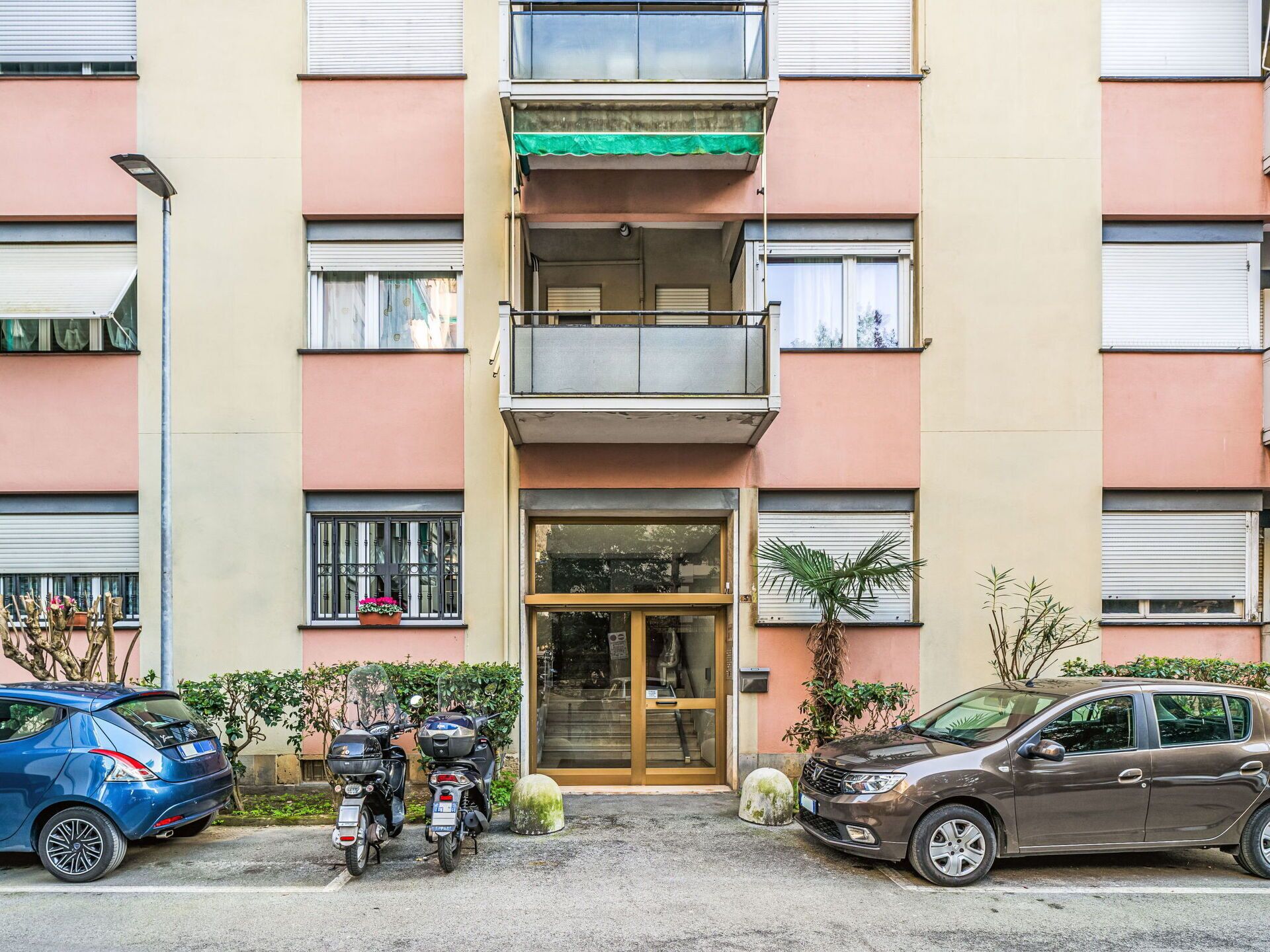 Un immeuble à plusieurs étages, avec une entrée au niveau du sol, des balcons et un espace de stationnement où sont garées des voitures et des motos.