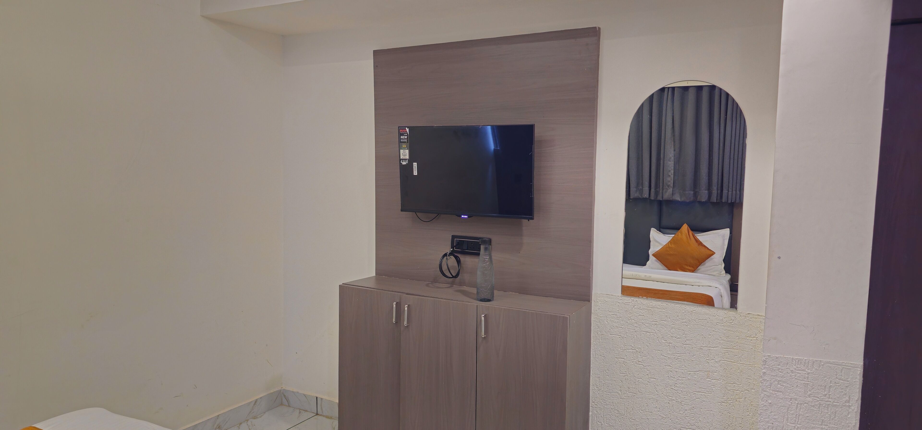 Une chambre d’hôtel équipée d’un téléviseur à écran plat fixé sur un panneau en bois au-dessus d’une commode, d’un lit avec une tête de lit et des oreillers, et d’une petite fenêtre avec des rideaux.