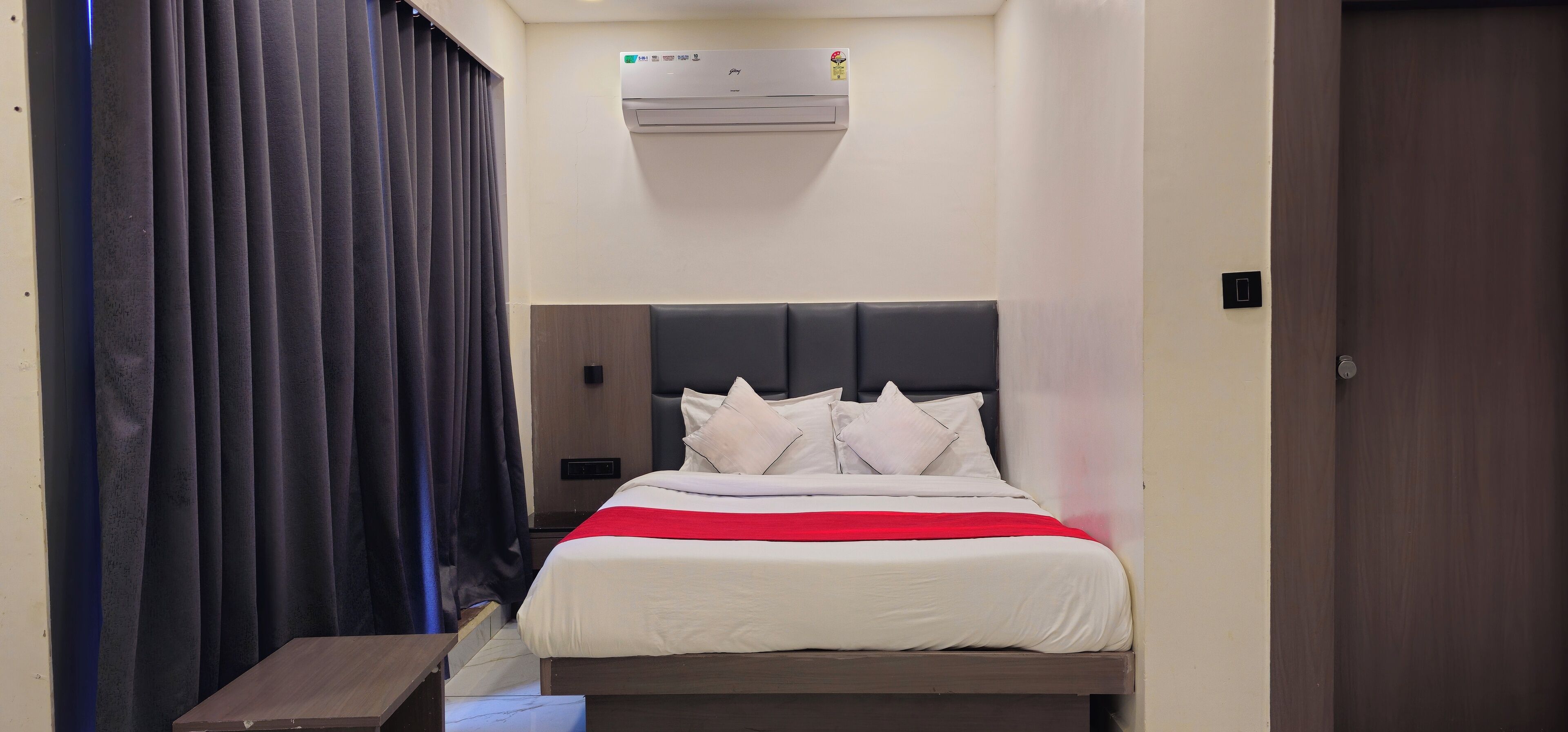Una habitación de hotel con cama, mesita de noche y aire acondicionado de pared, además de una puerta.