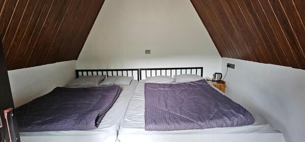 A room with two beds, wooden ceiling beams, white walls, and a small bedside table with a lamp. - The Soul of vattavada (Devikolam)