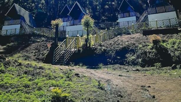 A row of cabins with a forest backdrop, a dirt path leading to them, and a clear sky. - The Soul of vattavada (Devikolam)