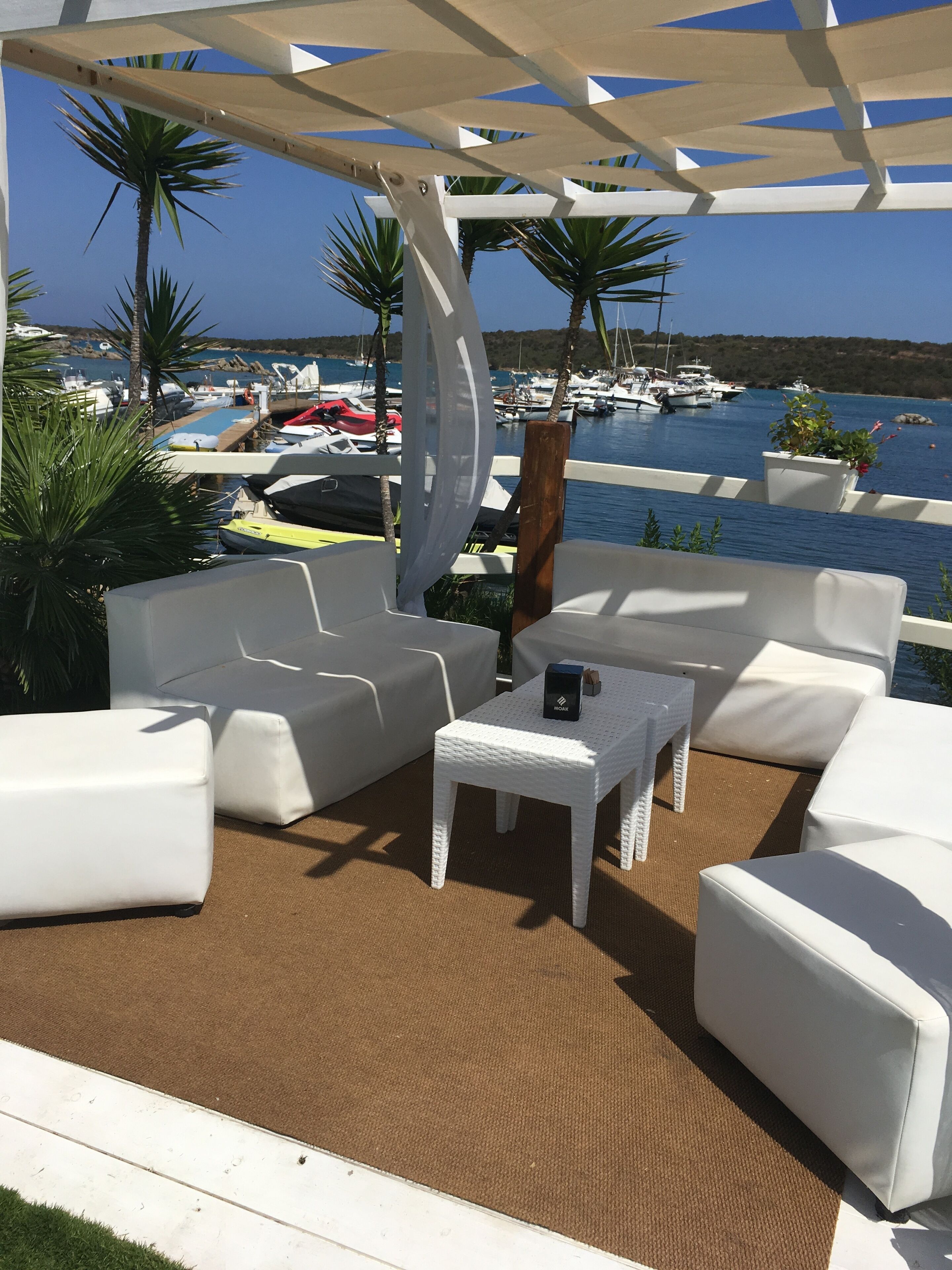Un espace extérieur aménagé avec des meubles blancs, une petite table et abrité, offrant une vue sur une marina avec des bateaux et des palmiers.