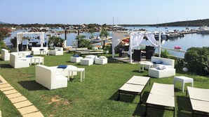 A seaside patio with white lounge chairs and tables, a clear view of the marina, and a grassy area. - THREE-ROOM APARTMENT HOUSE EMY C11 GROUND FLOOR WITH PATIO AND GARDEN (Golfo di Marinella)