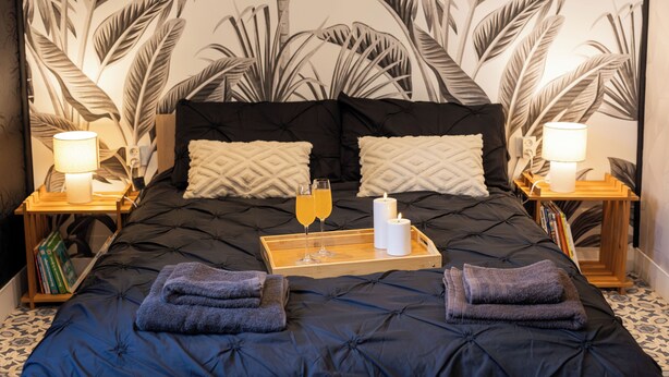 Un lit avec une couverture sombre, deux oreillers, un plateau avec deux verres de jus d’orange, deux bougies et des serviettes pliées.