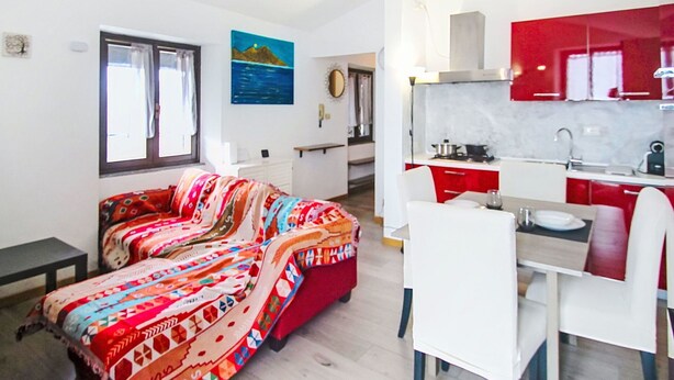 Ein modernes Wohnzimmer mit einem farbenfrohen Sofa, einem Essbereich mit Tisch und Stühlen sowie einer Küche mit roten Schränken und weißen Geräten.