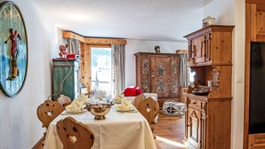 A dining area with a wooden table set for four, surrounded by wooden chairs. A large wooden cabinet with intricate carvings is to the right, and a green cabinet with a figure on top is in the background. - Beautiful apartment for 5 guests with TV, patio, pets allowed and panoramic view (Silvaplana)