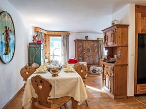 Un coin repas avec une table en bois dressée pour quatre personnes, entourée de chaises en bois. Un grand meuble en bois aux sculptures complexes se trouve à droite, et un meuble vert surmonté d’une statuette se distingue à l’arrière-plan.
