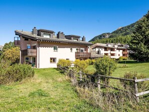A two-story building with a balcony, surrounded by a grassy area and a wooden fence. - Lovely apartment for 5 guests with TV, patio, pets allowed and panoramic view (Silvaplana)