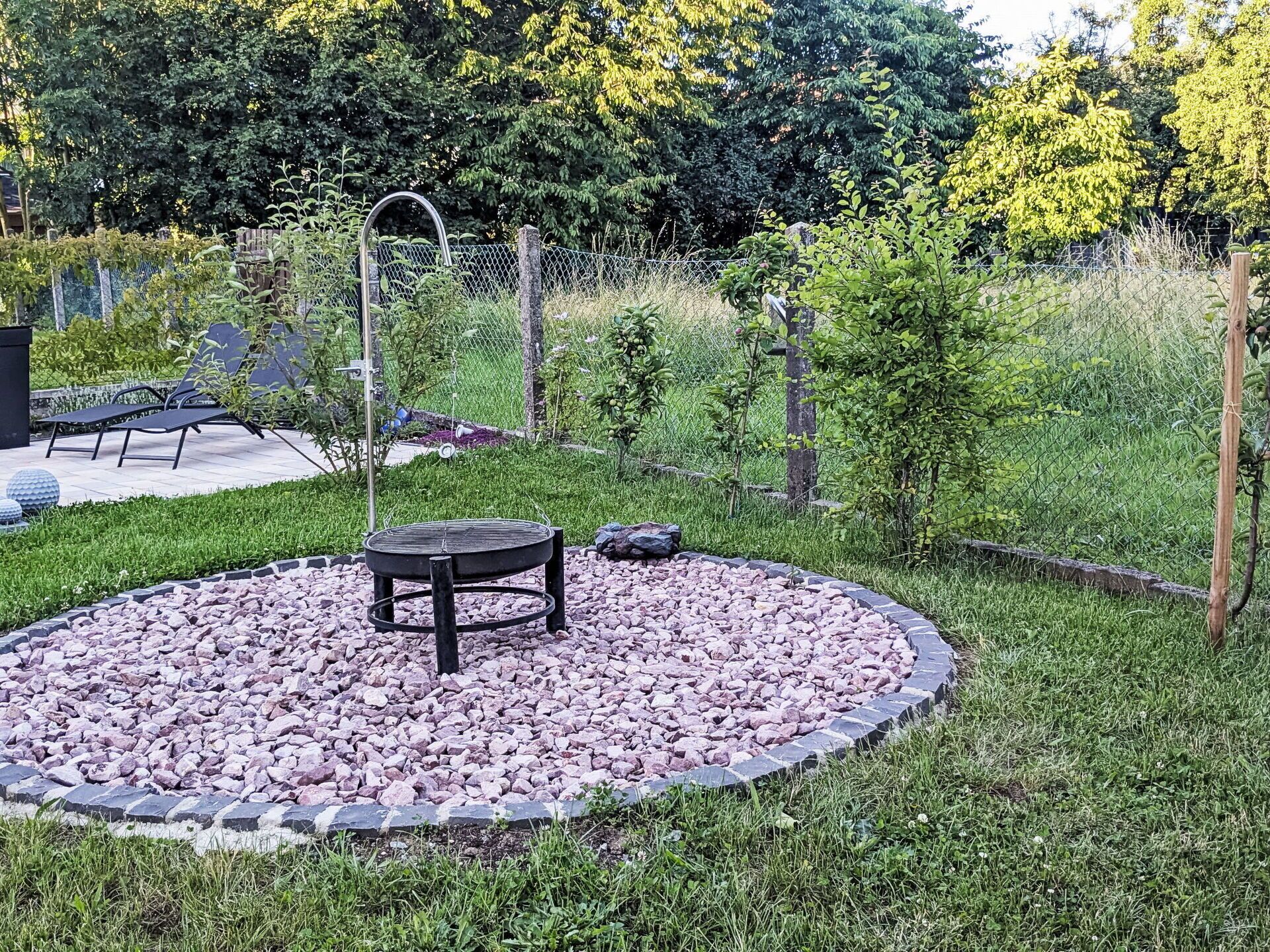 Un espacio circular para sentarse al aire libre, con una mesa y sillas de metal, rodeado por un borde de piedra y césped.