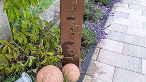 Ein Metallschild mit Schriftzug, umgeben von Grünpflanzen und einem gepflasterten Weg.