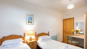 Une chambre avec deux lits simples, un bureau en bois avec une chaise, une table de chevet avec une lampe et une armoire en bois.