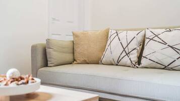A neutral-colored sofa with patterned cushions, a small wooden side table with a bowl of snacks, and a plain wall in the background.