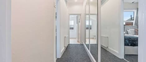 A hallway with a carpeted floor, a door leading to a room with a bed, and a large mirror reflecting the room.