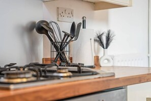 En köksbänk med en gasspis, ett förvaringsställ för köksredskap och en vit vas med dekorativa grenar.